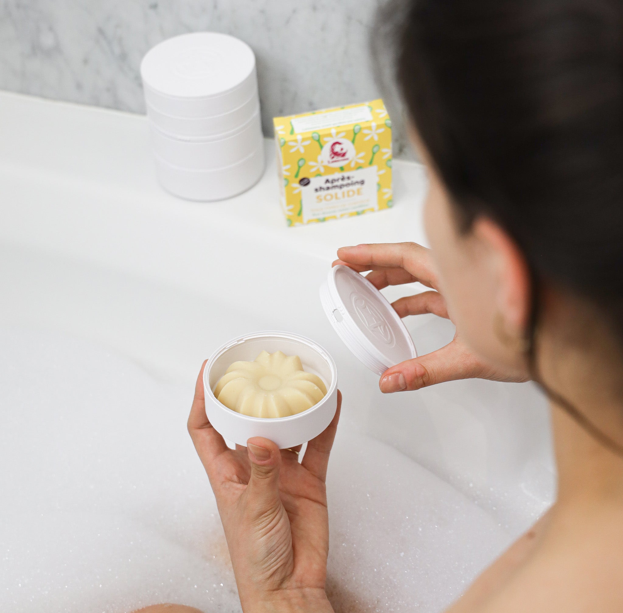 Après-shampoing de la marque Lamazuna, senteur vanille avec son packaging jaune dans les mains d'une personne dans un bain